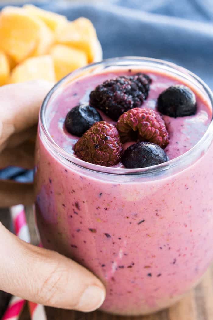 This Mango Berry Smoothie combines fresh mango with mixed berries into a refreshing smoothie recipe. This fruit smoothie is perfect for breakfast or a snack! a hand holding a glass filled with a Mango Berry Smoothie