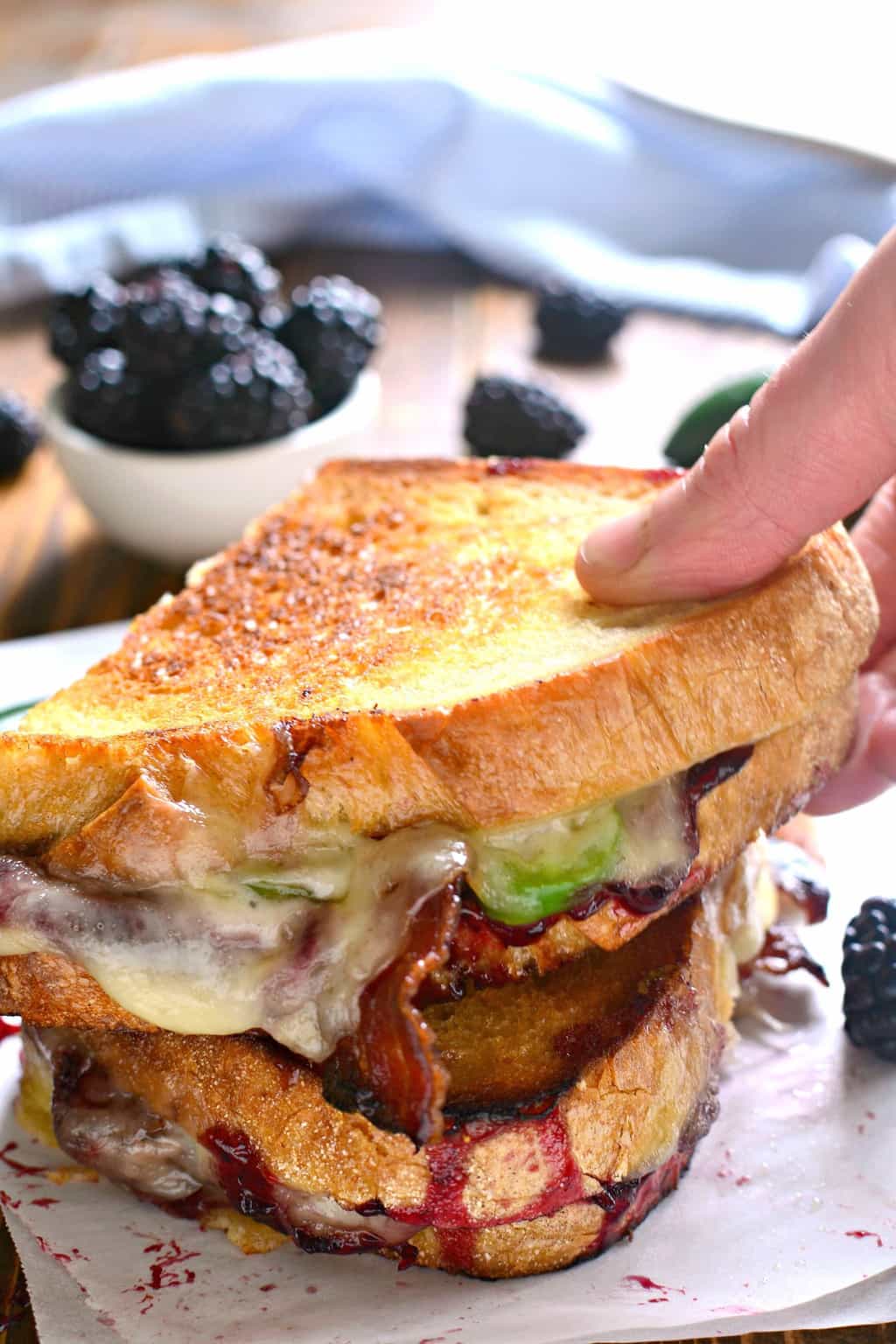 Blackberry Bacon Grilled Cheese is the perfect combination of savory and sweet! Made with Swiss cheese, blackberry jam, fresh jalapeños, and crispy bacon, it's a must try for ALL sandwich lovers! hand holding a Blackberry Bacon Grilled Cheese