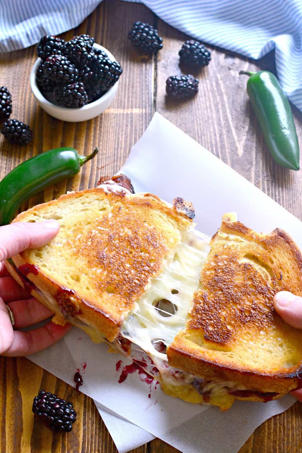 Blackberry Bacon Grilled Cheese is the perfect combination of savory and sweet! Made with Swiss cheese, blackberry jam, fresh jalapeños, and crispy bacon, it's a must try for ALL sandwich lovers! hands pulling apart a Blackberry Bacon Grilled Cheese so you can see the melted cheese stretching between the two halves