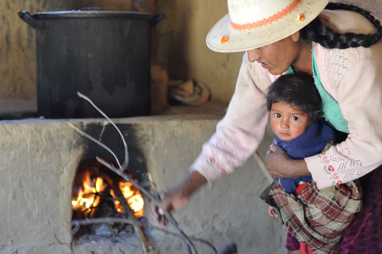 a Bolivian mother holding her baby as she stokes a wood burning stove
