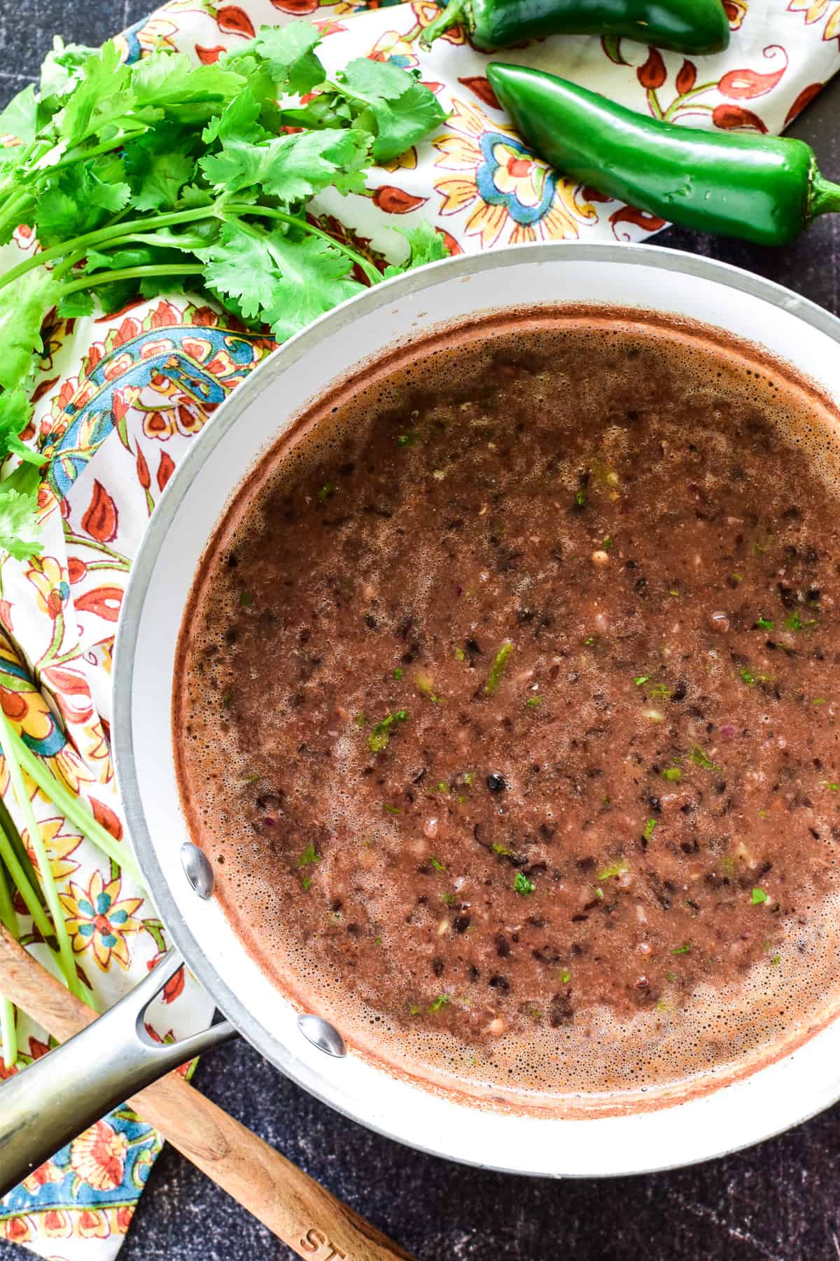 Overhead shot of Black Bean Soup in a white saucepan with a patterned towel and fresh garnishes