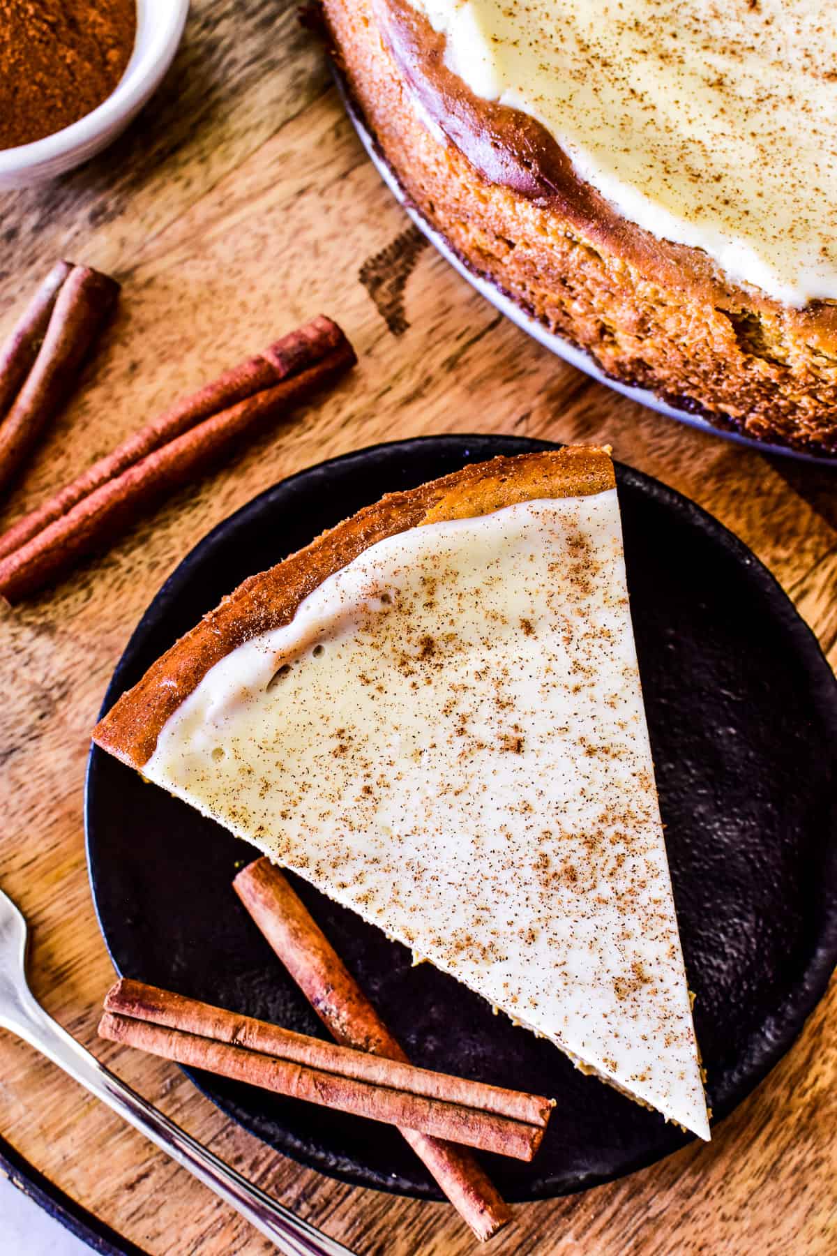 Overhead shot of a slice of pumpkin cheesecake on a dark plate with cinnamon sticks