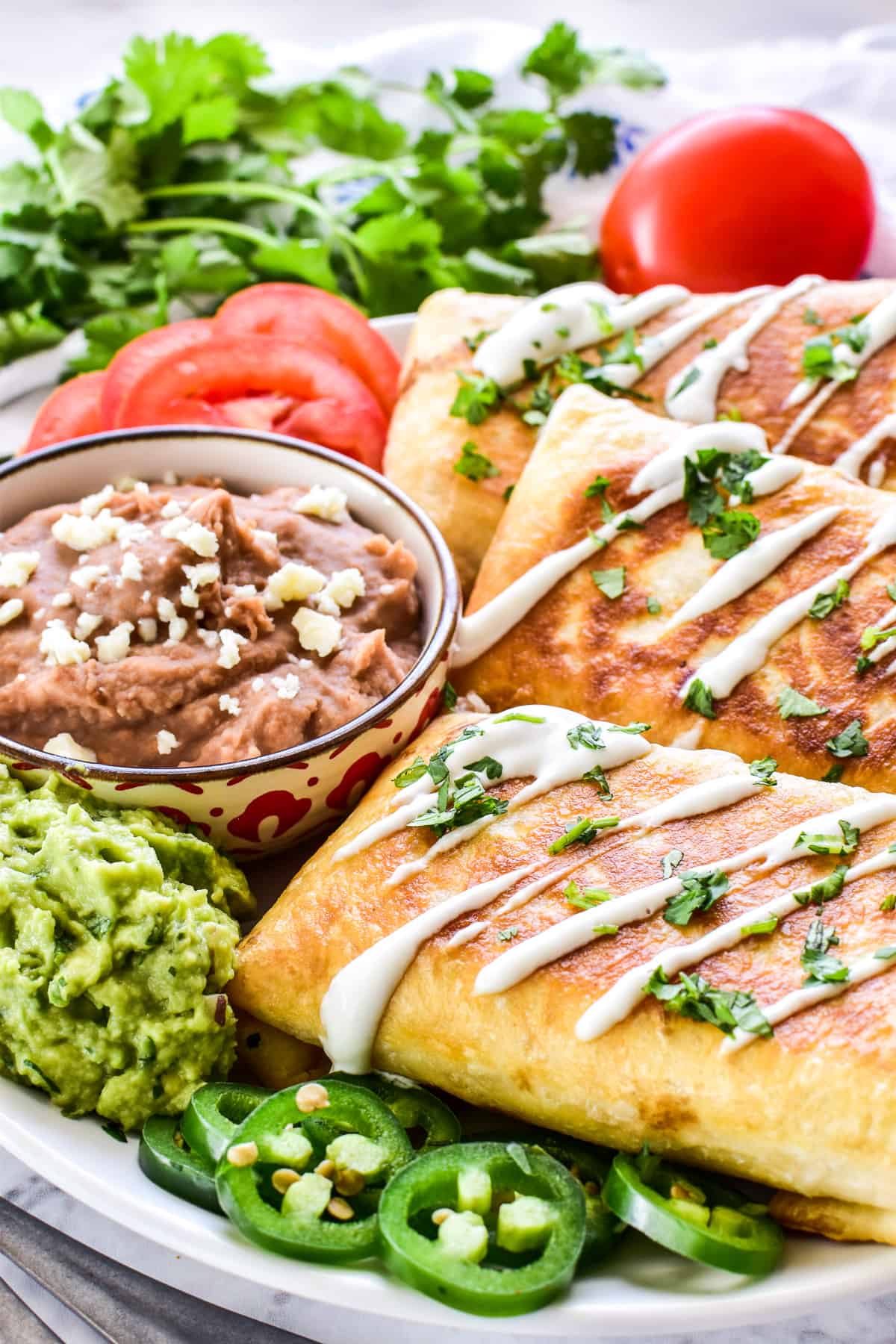 Chicken Chimichangas on a plate with refried beans, tomatoes, guacamole, and sliced jalapeño