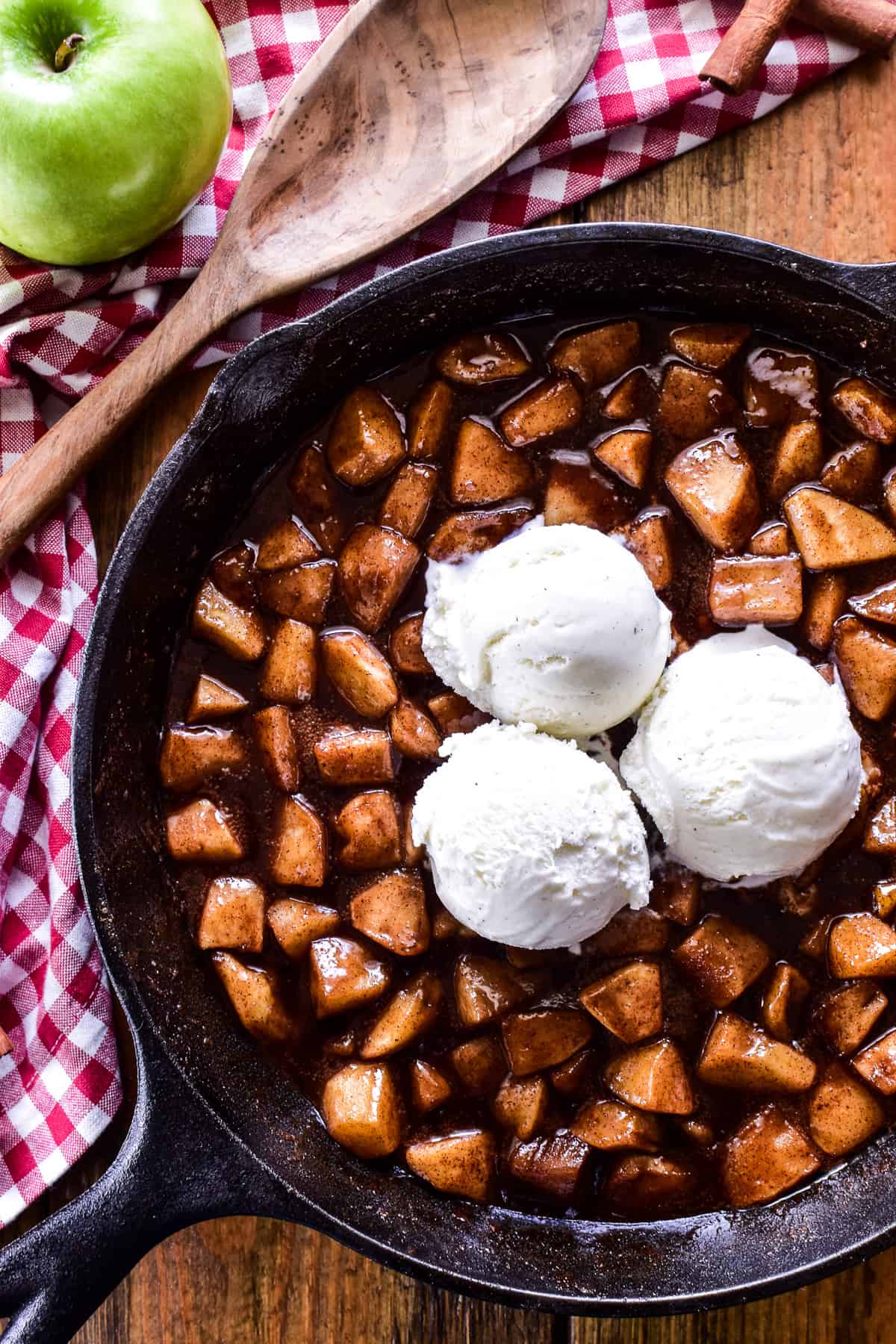 Fried Apples in a cast iron skillet with 3 scoops of vanilla ice cream and a wooden serving spoon