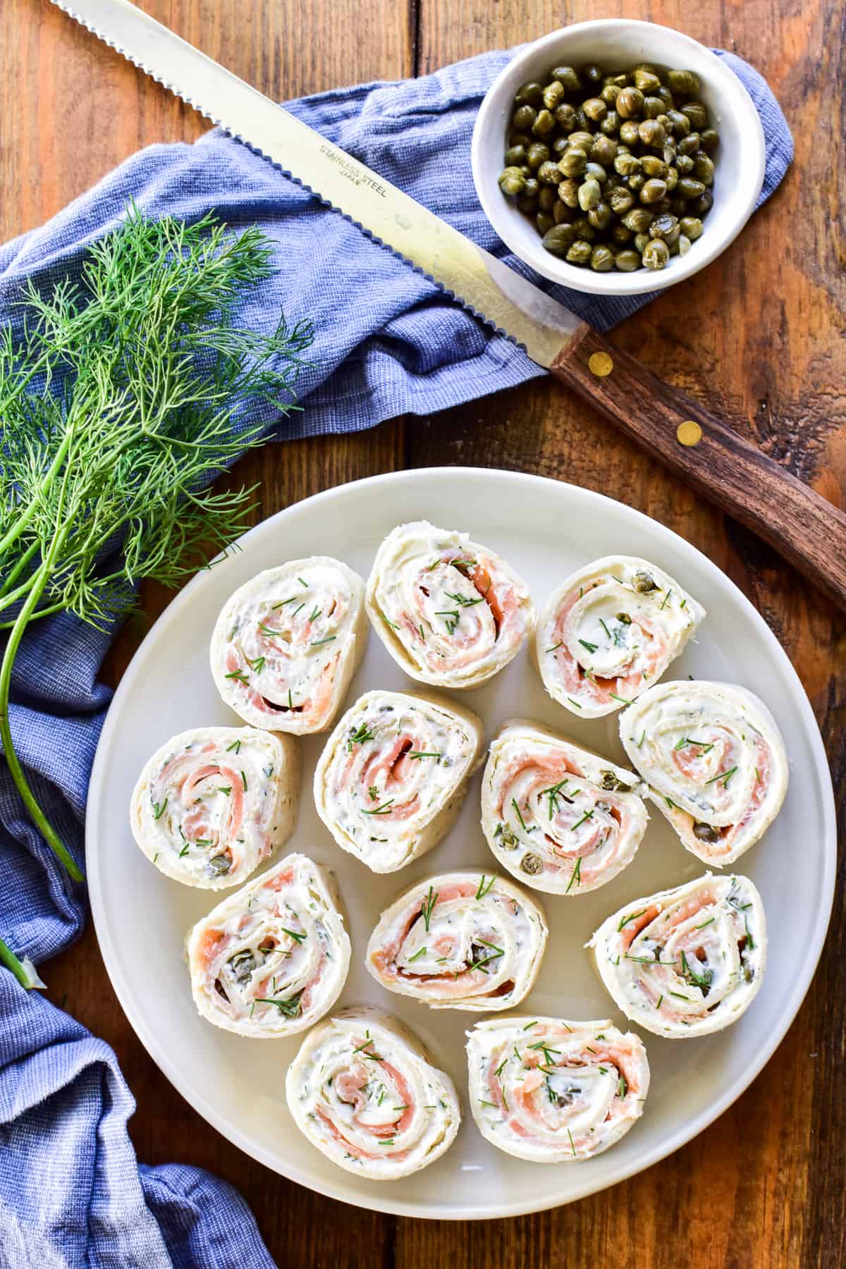 Overhead image of Lox Cream Cheese Pinwheels on a white plate with fresh dill and capers