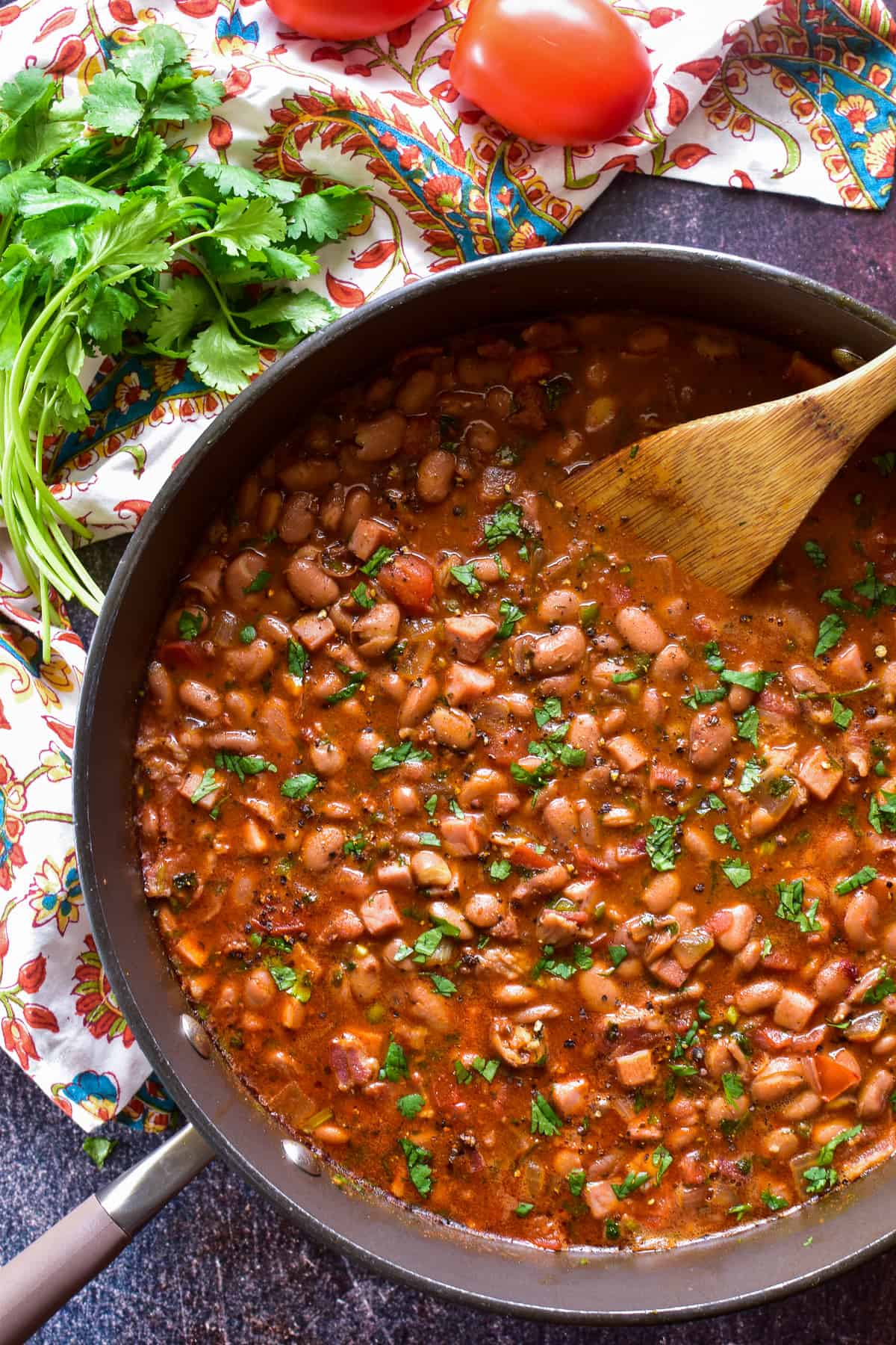 Mexican Charro Beans in a large skillet with fresh cilantro, tomatoes, and a patterned towel 